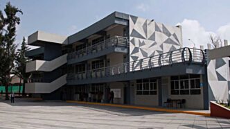 Modern grey building with external zigzag staircases and a geometric triangular mural on the side; people near the entrance on a paved plaza.
