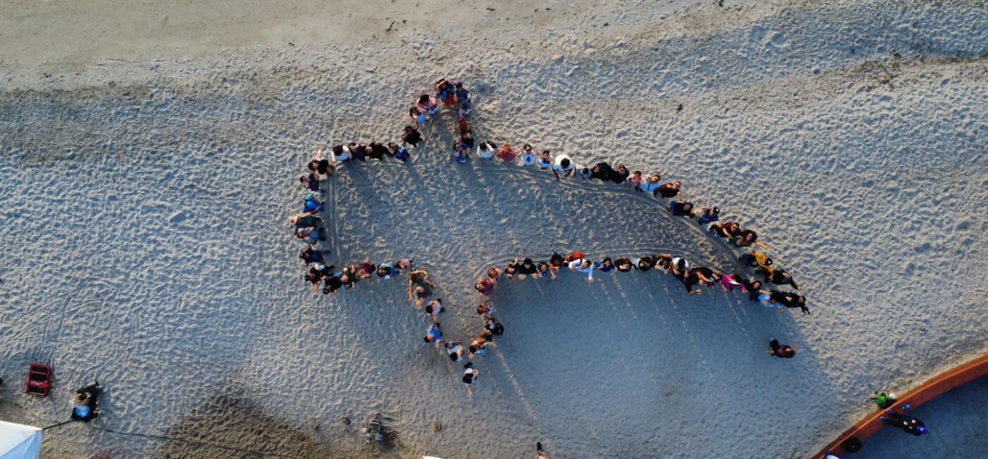 Aerial view of a group of people standing together on a sandy beach, forming a large star-shaped outline.