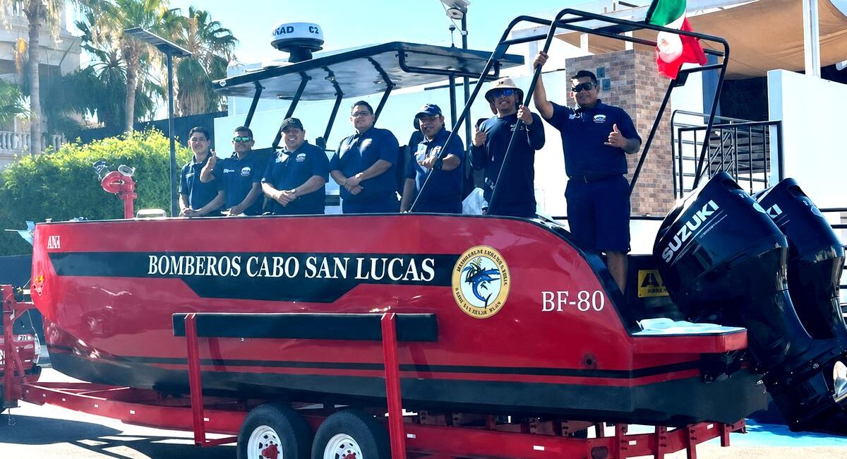 Red rescue boat on a trailer with six firefighters in navy shirts, Cabo San Lucas fire department, Mexico flag raised nearby.