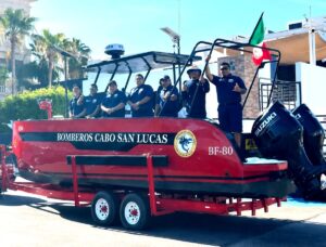 Red rescue boat on a trailer with six firefighters in navy shirts, Cabo San Lucas fire department, Mexico flag raised nearby.