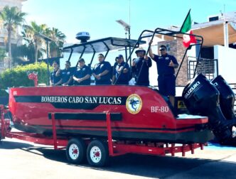 Red rescue boat on a trailer with six firefighters in navy shirts, Cabo San Lucas fire department, Mexico flag raised nearby.