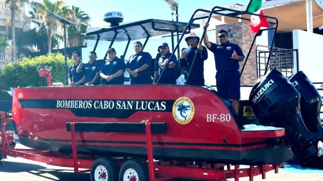 Red rescue boat on a trailer with six firefighters in navy shirts, Cabo San Lucas fire department, Mexico flag raised nearby.