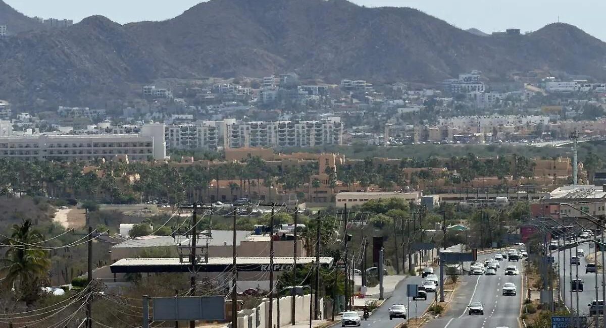 Wide city street scene with traffic and palm trees, set against a backdrop of low buildings and distant hills.