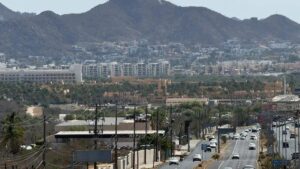 Wide city street scene with traffic and palm trees, set against a backdrop of low buildings and distant hills.