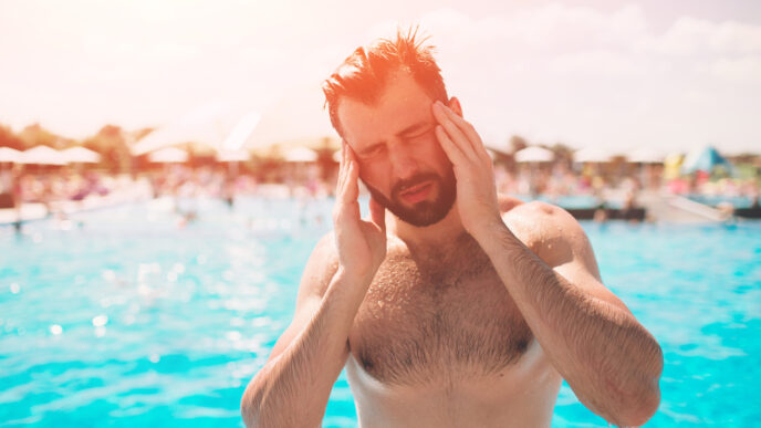 Bearded man at an outdoor pool, holding his temples with his eyes closed in the sun.