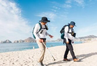 Two people in hats and backpacks walk along a sunny beach, one using a metal detector search for items in the sand.