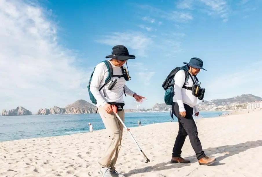 Beach-Audit - Gringo Gazette Two people in hats and backpacks walk along a sunny beach, one using a metal detector search for items in the sand.