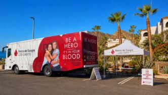 Blood donation truck with 'Be a Hero' message, and a tent for a blood bank outreach at a sunny parking lot in Los Cabos