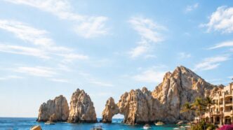 Beach scene in Los Cabos with rock formations, blue ocean, and people strolling along a promenade toward the water; sign shows Los Cabos, Baja California Sur.