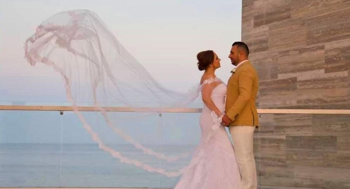 Bride in a white wedding dress with flowing veil stands with a groom in a tan jacket on a balcony overlooking the sea at sunset.