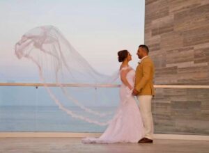 Bride in a white wedding dress with flowing veil stands with a groom in a tan jacket on a balcony overlooking the sea at sunset.