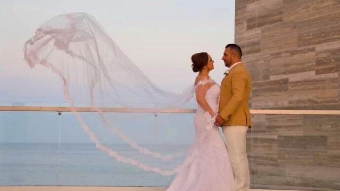 Bride in a white wedding dress with flowing veil stands with a groom in a tan jacket on a balcony overlooking the sea at sunset.