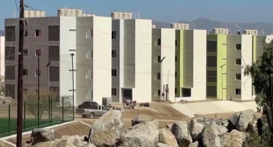 Row of modern white and light-green apartment buildings under construction, with a dirt street and boulders in the foreground.