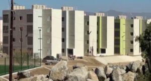 Row of modern white and light-green apartment buildings under construction, with a dirt street and boulders in the foreground.