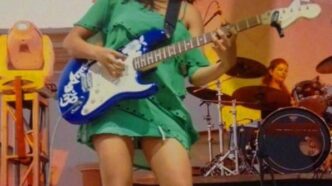 Female guitarist in a green dress playing a blue electric guitar on stage, with a drummer in the background.