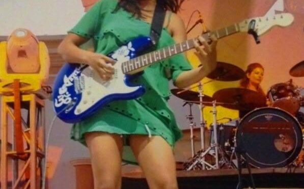 Female guitarist in a green dress playing a blue electric guitar on stage, with a drummer in the background.