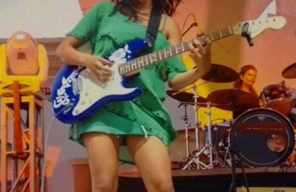 Female guitarist in a green dress playing a blue electric guitar on stage, with a drummer in the background.