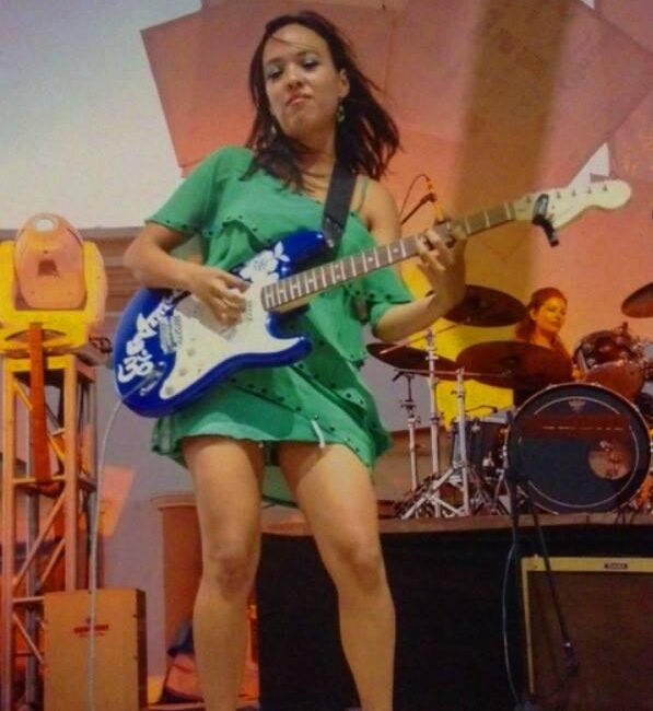 Female guitarist in a green dress playing a blue electric guitar on stage, with a drummer in the background.