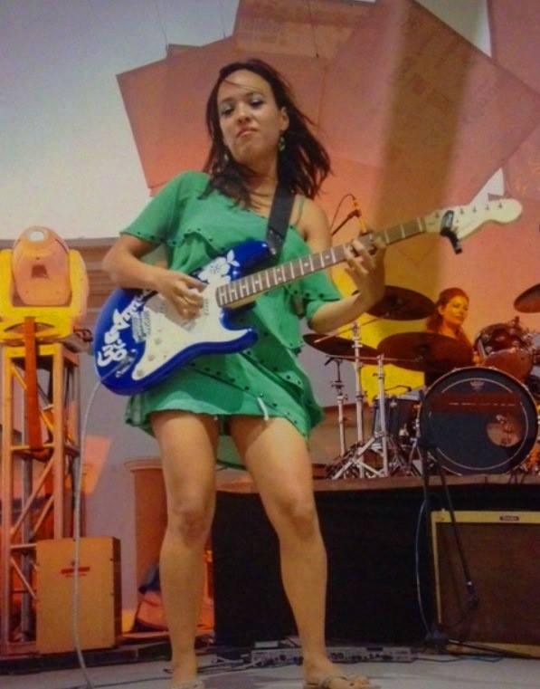 Female guitarist in a green dress playing a blue electric guitar on stage, with a drummer in the background.