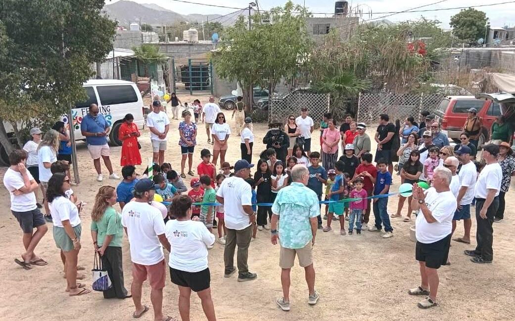 A diverse group of children and adults form a circle outdoors for a community event, with a blue ribbon across the center and onlookers surrounding them in a dusty yard.