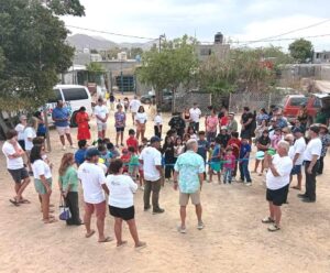 A diverse group of children and adults form a circle outdoors for a community event, with a blue ribbon across the center and onlookers surrounding them in a dusty yard.