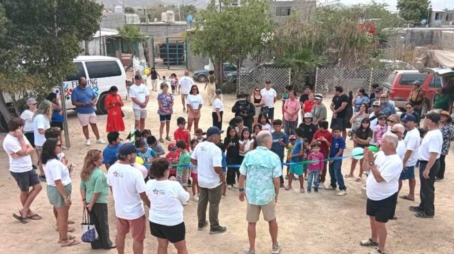 A diverse group of children and adults form a circle outdoors for a community event, with a blue ribbon across the center and onlookers surrounding them in a dusty yard.