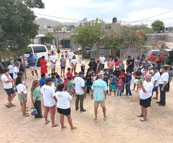 A diverse group of children and adults form a circle outdoors for a community event, with a blue ribbon across the center and onlookers surrounding them in a dusty yard.