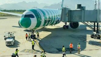 Green and white passenger airplane connected to a jet bridge at the gate with ground crew directing operations on the tarmac