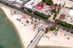 Aerial view of a sandy beach with clear green water, palm-lined promenade, and a row of white buildings along a street.