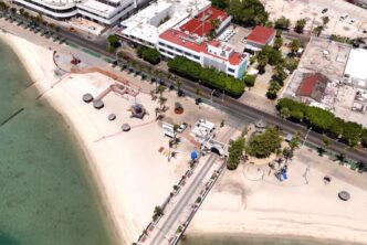 Aerial view of a sandy beach with clear green water, palm-lined promenade, and a row of white buildings along a street.