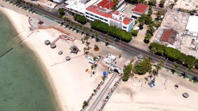 Aerial view of a sandy beach with clear green water, palm-lined promenade, and a row of white buildings along a street.