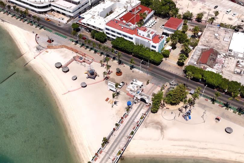 Aerial view of a sandy beach with clear green water, palm-lined promenade, and a row of white buildings along a street.