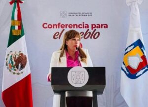 Mujer habla detrás de un podio en una conferencia gubernamental, con la bandera de México a la izquierda y otra bandera regional a la derecha