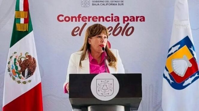 Mujer habla detrás de un podio en una conferencia gubernamental, con la bandera de México a la izquierda y otra bandera regional a la derecha