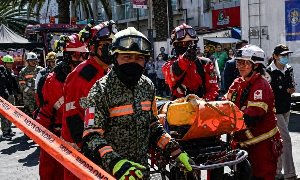 National-drill - Gringo Gazette Group of firefighters in red and camouflage uniforms carry a wheeled stretcher with an orange medical bag through a city street as bystanders watch.