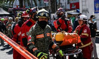 Group of firefighters in red and camouflage uniforms carry a wheeled stretcher with an orange medical bag through a city street as bystanders watch.