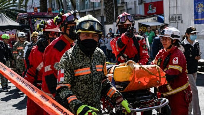 Group of firefighters in red and camouflage uniforms carry a wheeled stretcher with an orange medical bag through a city street as bystanders watch.
