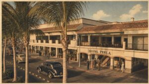 Wide street view of Hotel Perla, a two-story Mediterranean-style building with palm trees, vintage cars, and guests outside on a sunny day.