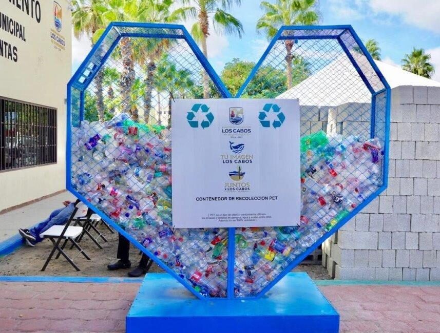 Heart-shaped blue recycling container filled with plastic bottles on a sunny outdoor plaza, with a sign on the front.