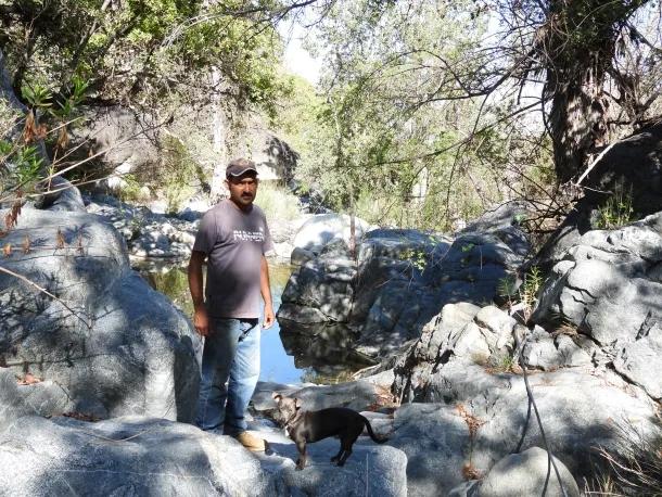Man in a gray t-shirt and jeans standing on large rocks beside a shallow creek, with a dog nearby among the boulders in a rocky canyon.