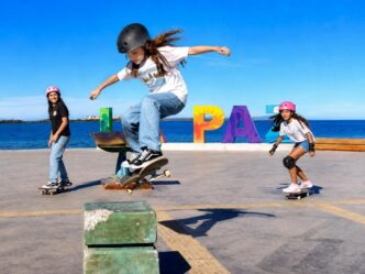 Three kids skateboarding at a seaside skatepark, one jumping a ramp while wearing a helmet; colorful PLAY letters in the background.