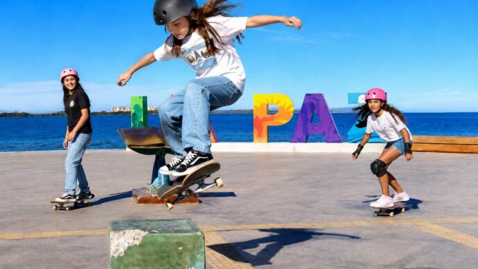 Three kids skateboarding at a seaside skatepark, one jumping a ramp while wearing a helmet; colorful PLAY letters in the background.