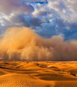 Desert dunes under a dramatic sky with a large dust cloud rolling over the horizon at sunset.