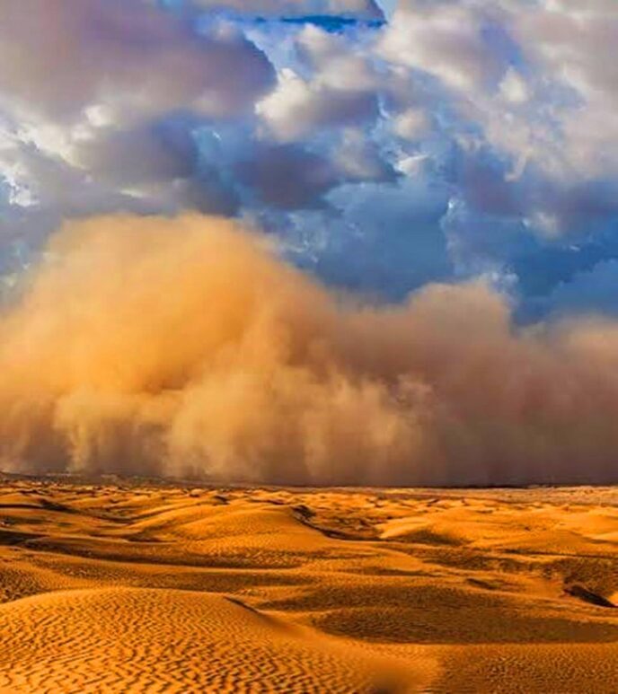 Desert dunes under a dramatic sky with a large dust cloud rolling over the horizon at sunset.