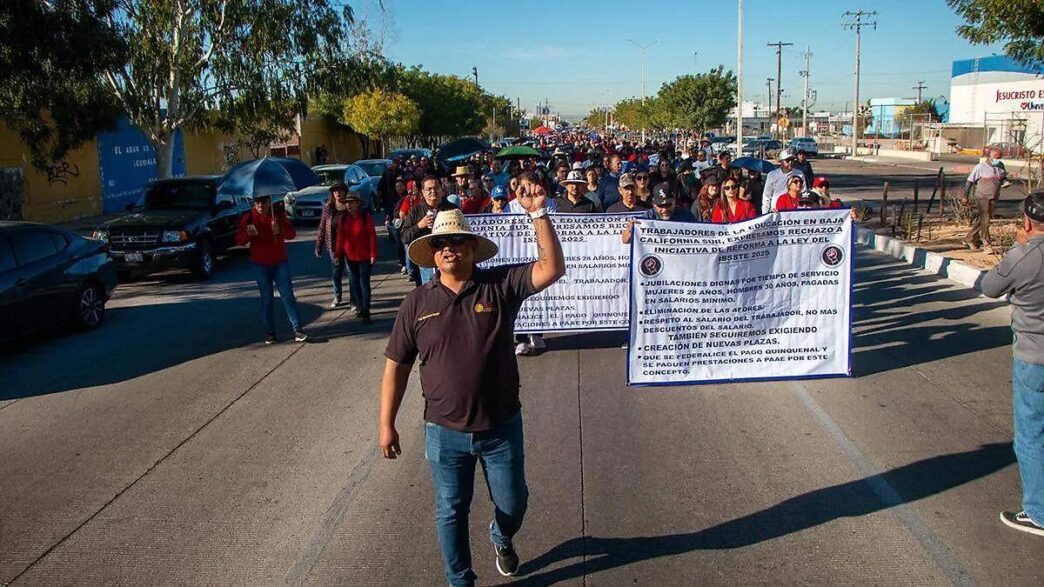 A man wearing a straw hat waves at the camera as he leads a street protest with a large banner and a crowd behind him.