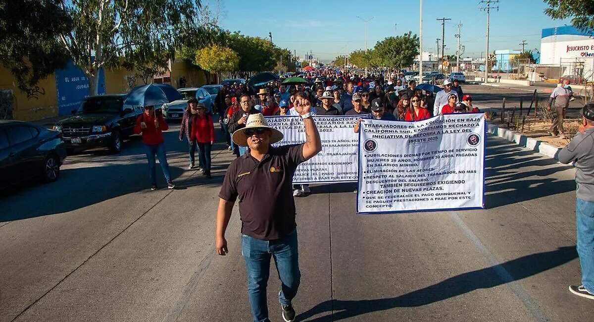 A man wearing a straw hat waves at the camera as he leads a street protest with a large banner and a crowd behind him.