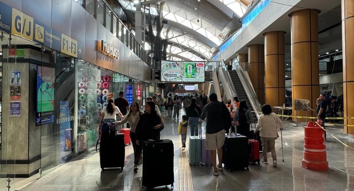 Crowded airport terminal with passengers pulling rolling suitcases, storefronts on the left and escalators ahead.