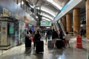 Crowded airport terminal with passengers pulling rolling suitcases, storefronts on the left and escalators ahead.