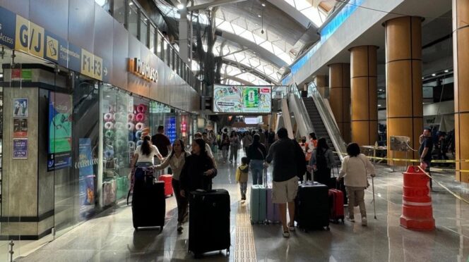 Crowded airport terminal with passengers pulling rolling suitcases, storefronts on the left and escalators ahead.