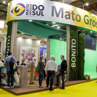 Trade show booth for Mato Grosso do Sul at a convention; visitors gather near a reception desk under a curved banner that reads 'Mato Grosso do Sul', with green vertical columns labeled 'Bonito' surrounding the space.